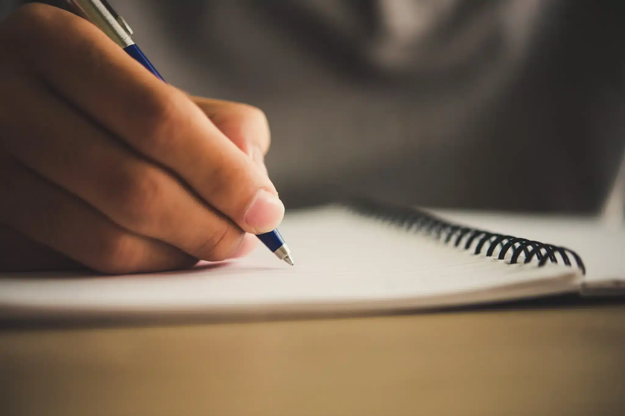 Pessoa escrevendo em caderno espiral com caneta azul, representando a redação dissertativa-argumentativa exigida em provas de língua portuguesa.