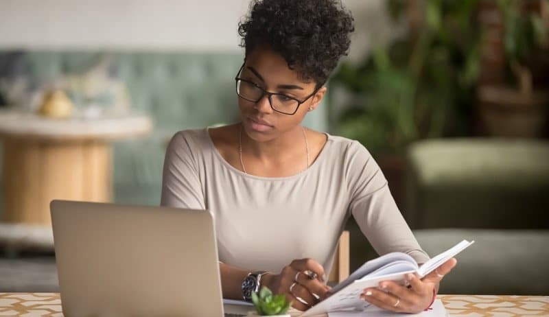Jovem mulher negra, usando óculos, concentrada em estudo à distância, fazendo anotações em um caderno ao lado de um notebook, focando na aquisição de novas competências para o mercado.
