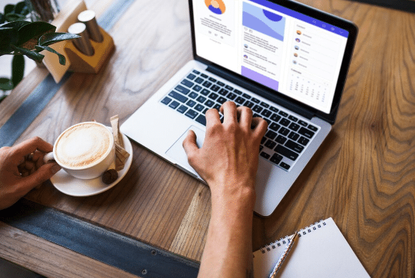 Pessoa usando laptop com gráficos na tela e segurando xícara de café em mesa de madeira