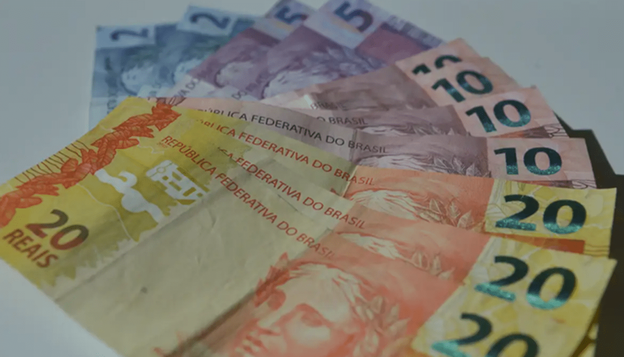 Notas de dinheiro brasileiras em várias denominações de cinco, dez e vinte reais espalhadas sobre superfície branca.
