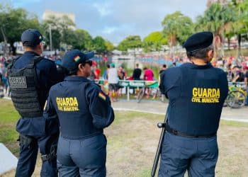 Três agentes da Guarda Civil Municipal de costas, vigiando multidão em parque com lago e pessoas ao fundo.