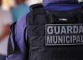 Vista traseira de guarda municipal com colete tático preto e uniforme azul em ambiente desfocado.