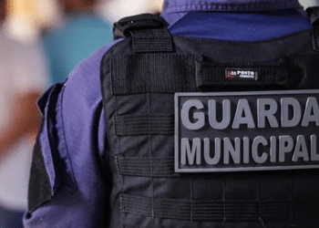 Vista traseira de guarda municipal com colete tático preto e uniforme azul em ambiente desfocado.