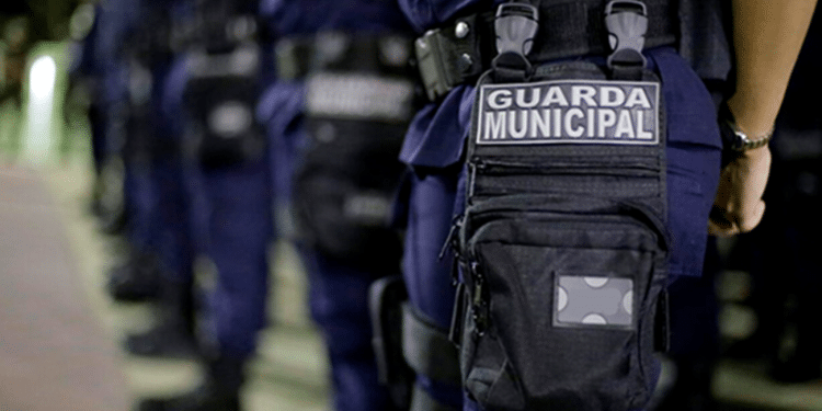 Guarda Municipal alinhada em fila, com uniformes azuis e bolsas táticas, em ambiente externo à noite.