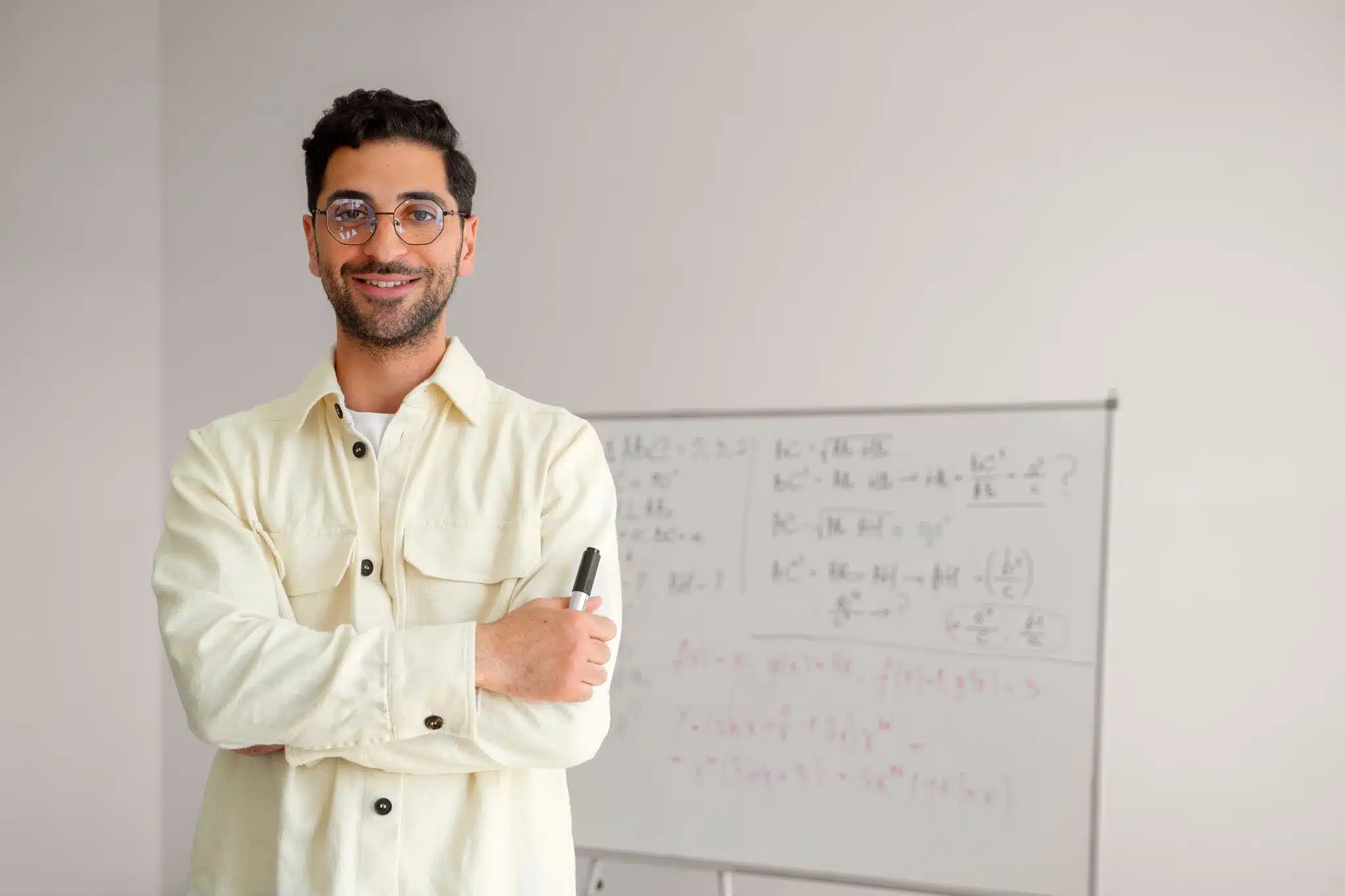 Homem jovem usando óculos e camisa clara com marcador na mão, ao lado de quadro branco com equações matemáticas