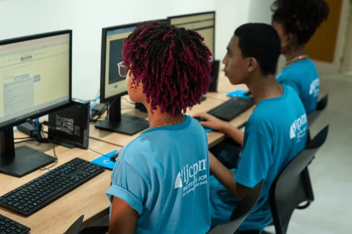 Três jovens sentados em cadeiras usando computadores com monitores em uma sala de aula.