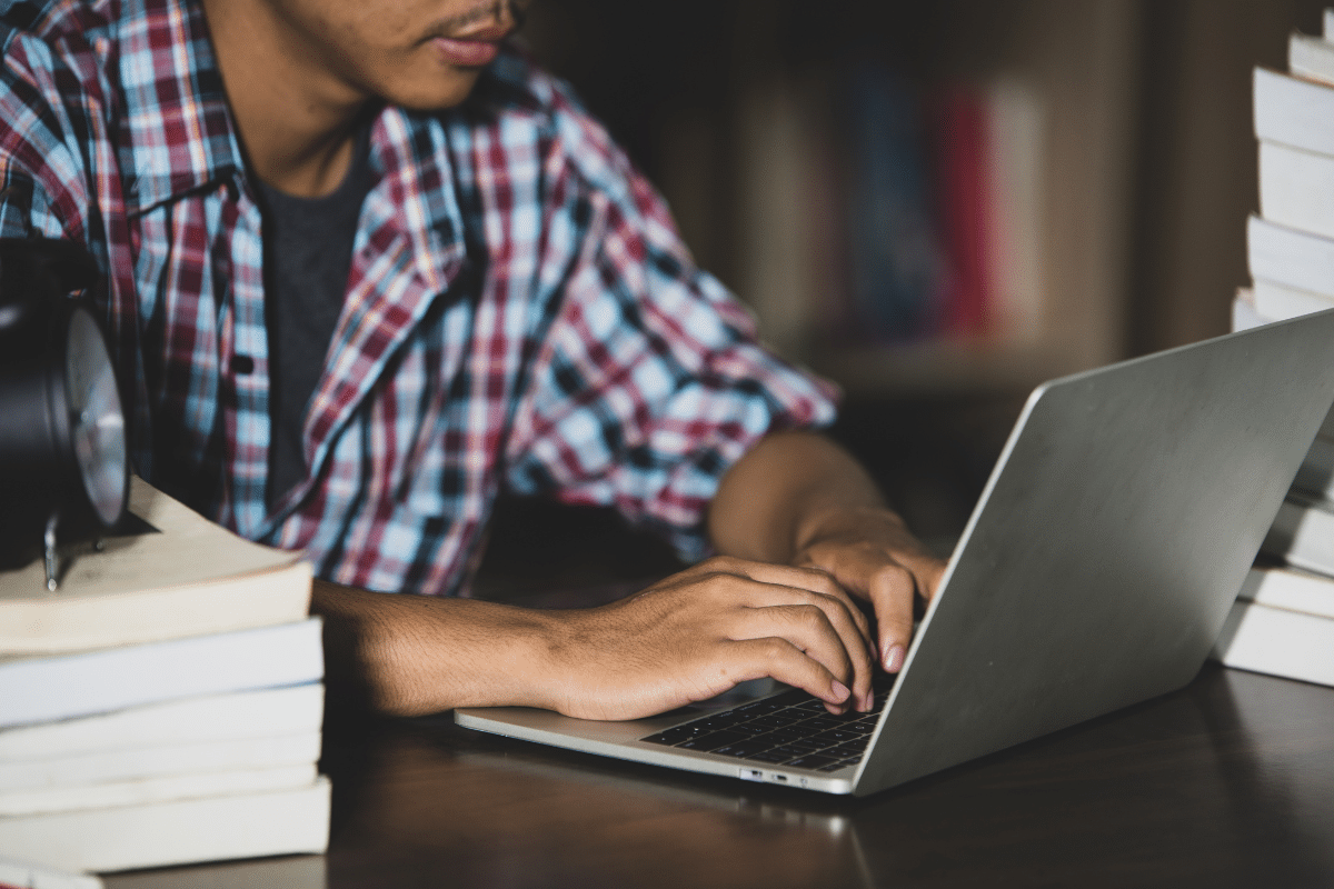 Pessoa digitando no laptop cercada por livros empilhados em mesa de estudo.