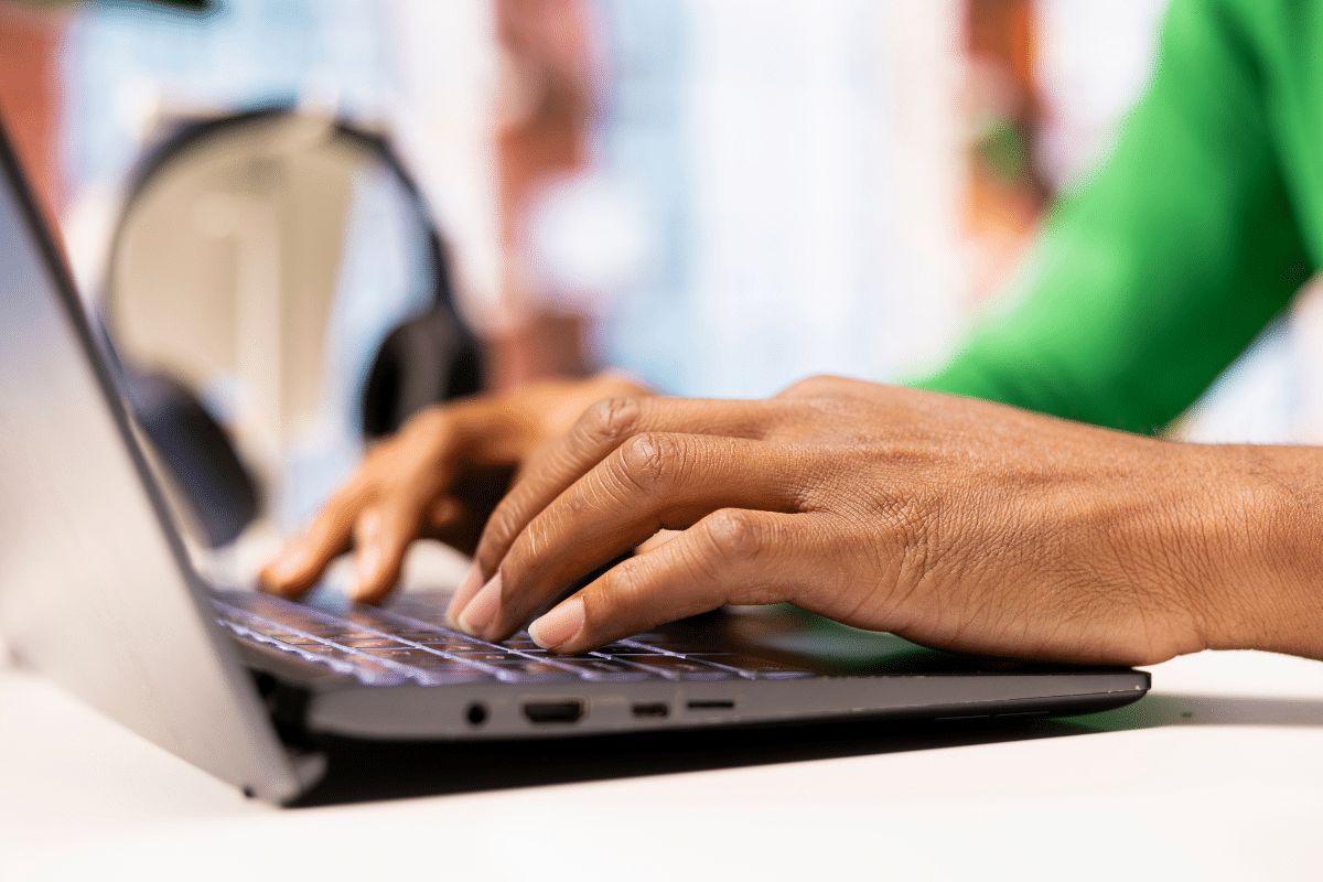 Mãos de pessoa negra digitando em teclado de laptop cinza sobre mesa branca