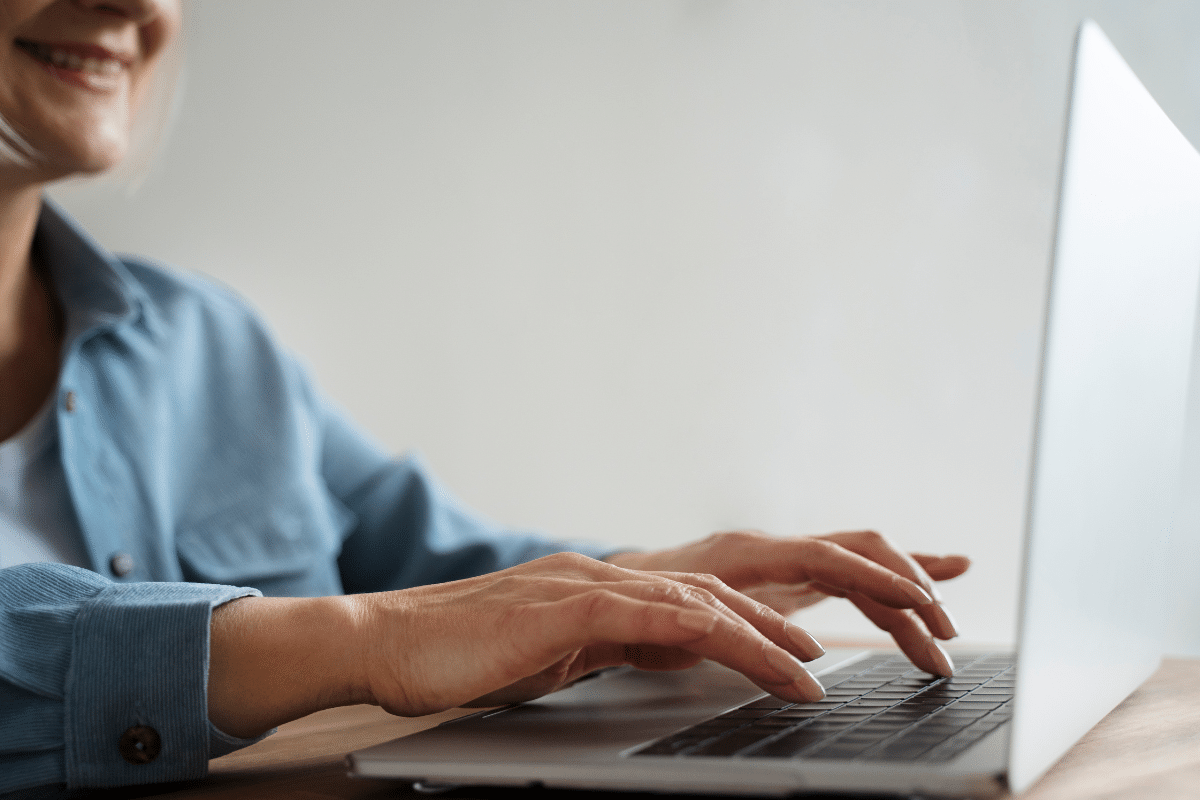 Mulher sorrindo enquanto digita em um laptop prata com camisa azul em fundo branco