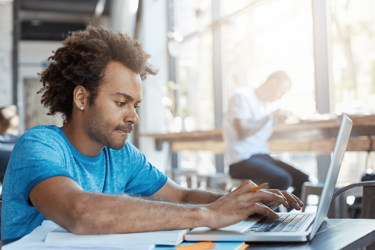 Homem jovem com cabelo cacheado usando camiseta azul, estudando em laptop dentro de café iluminado