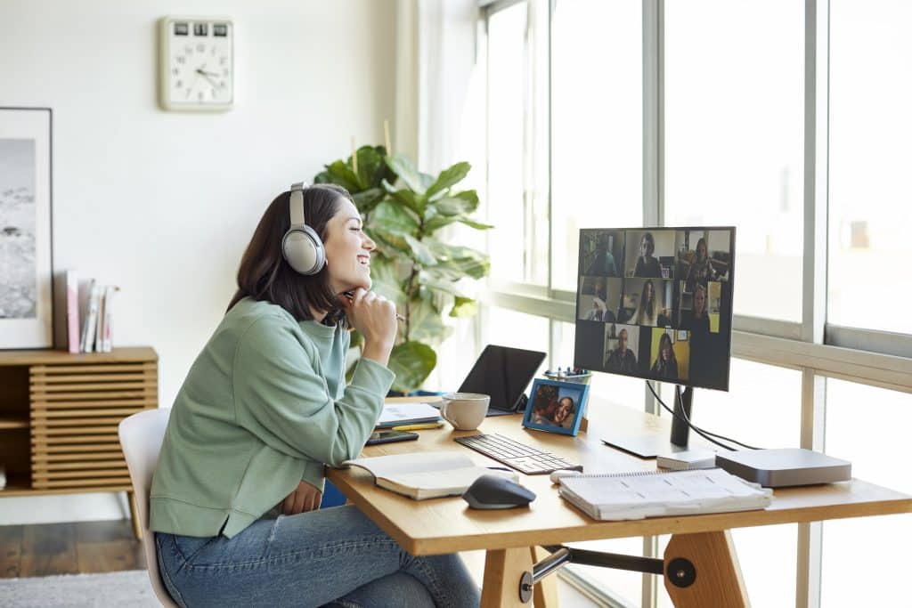 Mulher participando de uma videoconferência em home office, com fones de ouvido e sorriso no rosto, enquanto interage com seus colegas no computador.