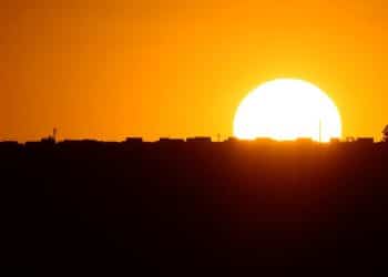 Um pôr do sol alaranjado sobre a silhueta de uma cidade, simbolizando as mudanças de horário do verão.