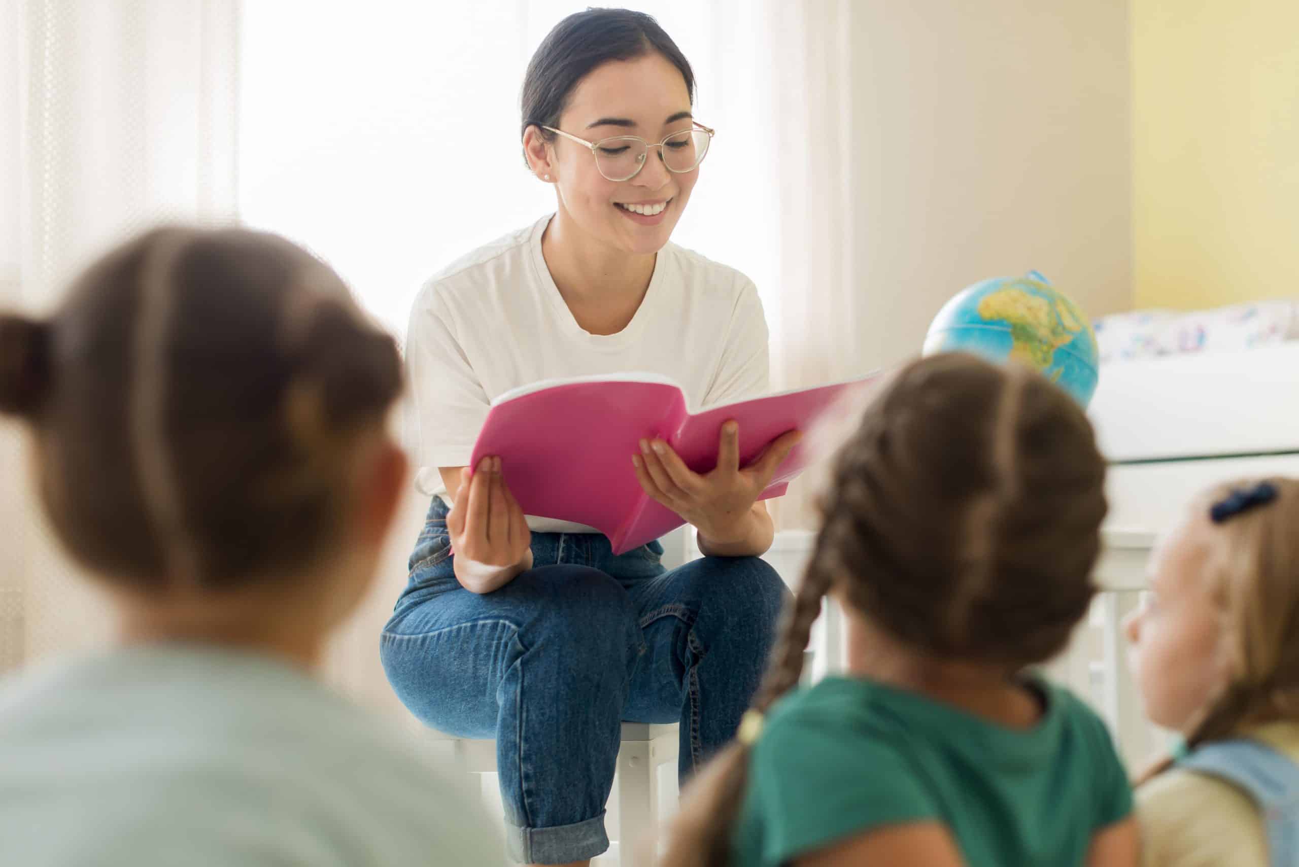 Professora asiática sorridente lendo um livro para um grupo de crianças pequenas em sala de aula. A imagem ilustra a atuação na educação pública para formados em Pedagogia, um dos cursos para sucesso na carreira pública.