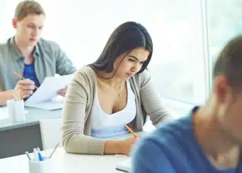 Jovem concentrada realizando prova de concurso em sala de aula com outros candidatos ao fundo em ambiente de seleção pública