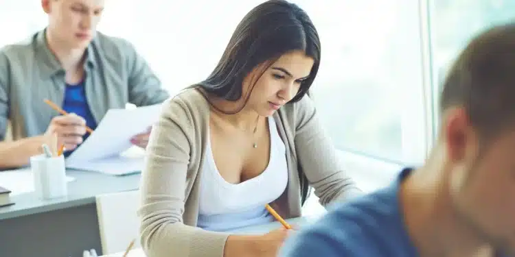 Jovem concentrada realizando prova de concurso em sala de aula com outros candidatos ao fundo em ambiente de seleção pública