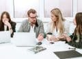 Grupo de quatro colegas de trabalho, com um homem gesticulando no centro, em uma reunião de brainstorming ou discussão, representando o momento da negociação e diálogo de ideias.