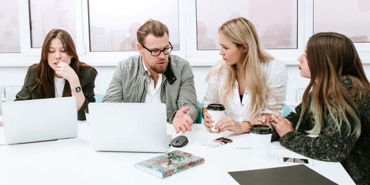Grupo de quatro colegas de trabalho, com um homem gesticulando no centro, em uma reunião de brainstorming ou discussão, representando o momento da negociação e diálogo de ideias.