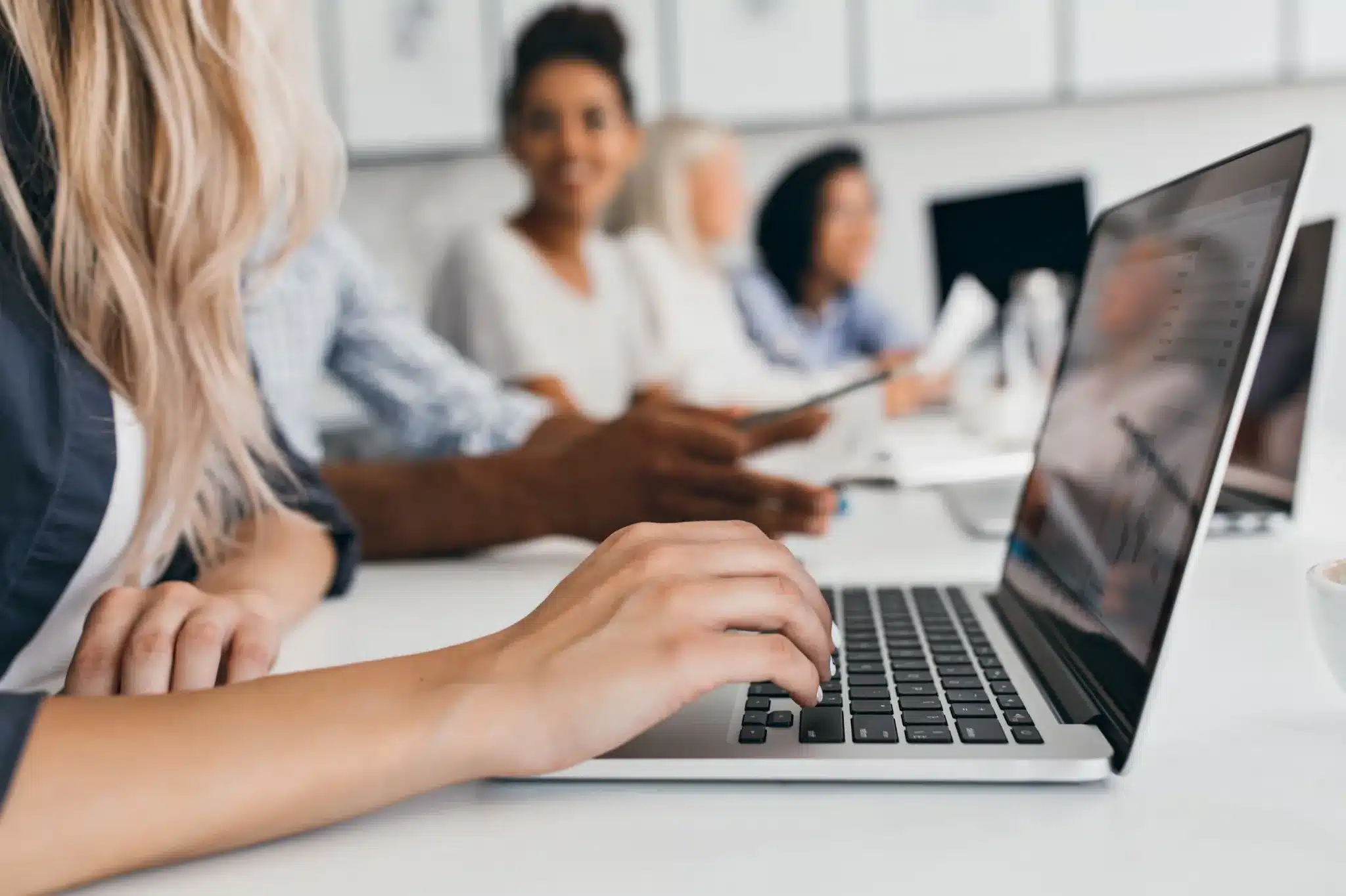 Mãos de pessoa digitando em laptop durante reunião de equipe com colegas ao fundo em escritório moderno