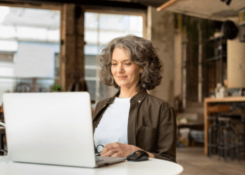 Mulher com cabelo grisalho sentada em mesa branca usando laptop em ambiente moderno e iluminado