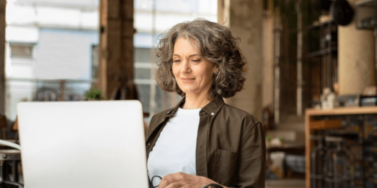 Mulher com cabelo grisalho sentada em mesa branca usando laptop em ambiente moderno e iluminado