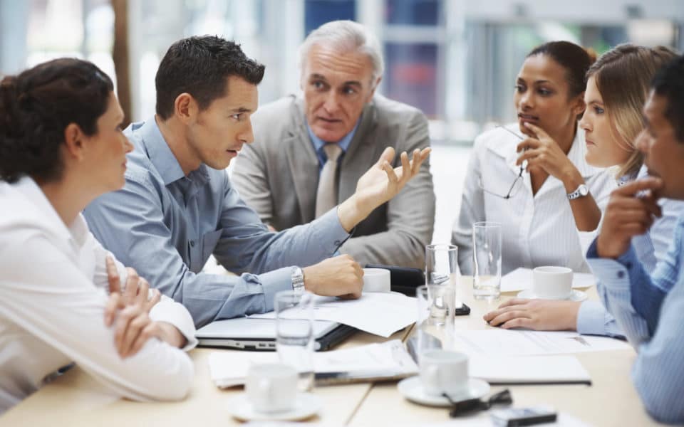 Grupo diverso de profissionais em uma mesa de reunião formal, gesticulando e debatendo, ilustrando a negociação e a comunicação estratégica no ambiente de trabalho.