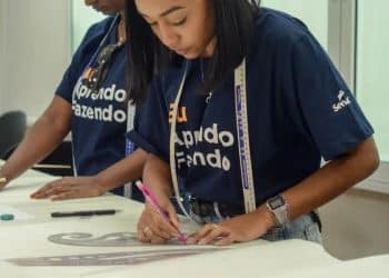 Duas alunas do Senac em uma aula prática, uma delas desenhando moldes em tecido, simbolizando o aprendizado técnico e profissionalizante.