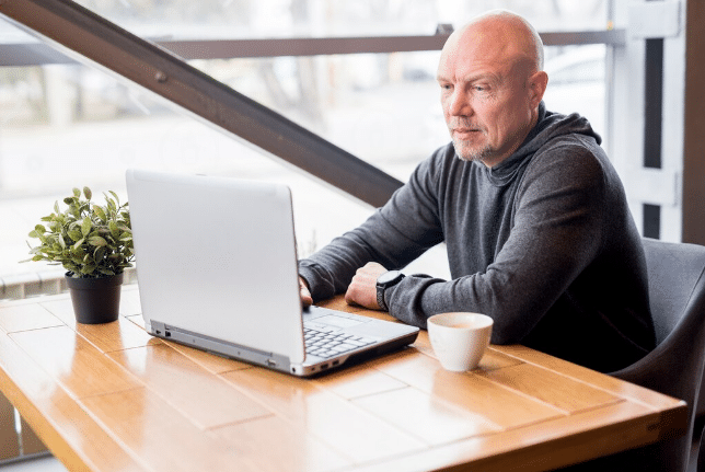 Homem calvo de meia-idade usando suéter cinza, sentado em cafeteria, trabalhando em laptop com xícara de café ao lado