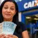 Mulher segura notas de real em frente a uma agência da Caixa, representando a solicitação do saque de R$ 6.220 do FGTS até 23 de julho.