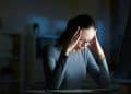 Mulher com óculos e expressão de cansaço ou estresse, segurando a cabeça em um ambiente escuro com a luz de um monitor, simbolizando sobrecarga mental e ansiedade.