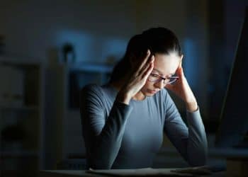 Mulher com óculos e expressão de cansaço ou estresse, segurando a cabeça em um ambiente escuro com a luz de um monitor, simbolizando sobrecarga mental e ansiedade.