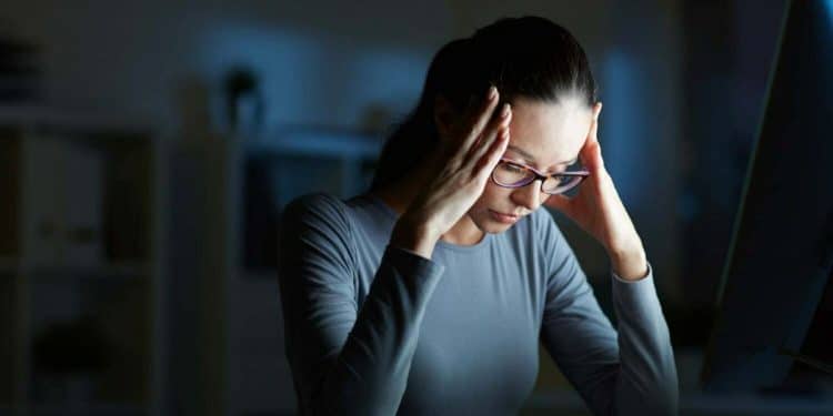Mulher com óculos e expressão de cansaço ou estresse, segurando a cabeça em um ambiente escuro com a luz de um monitor, simbolizando sobrecarga mental e ansiedade.