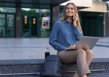 Mulher jovem sorridente sentada ao ar livre com laptop no colo e falando ao celular, mochila e garrafa ao lado
