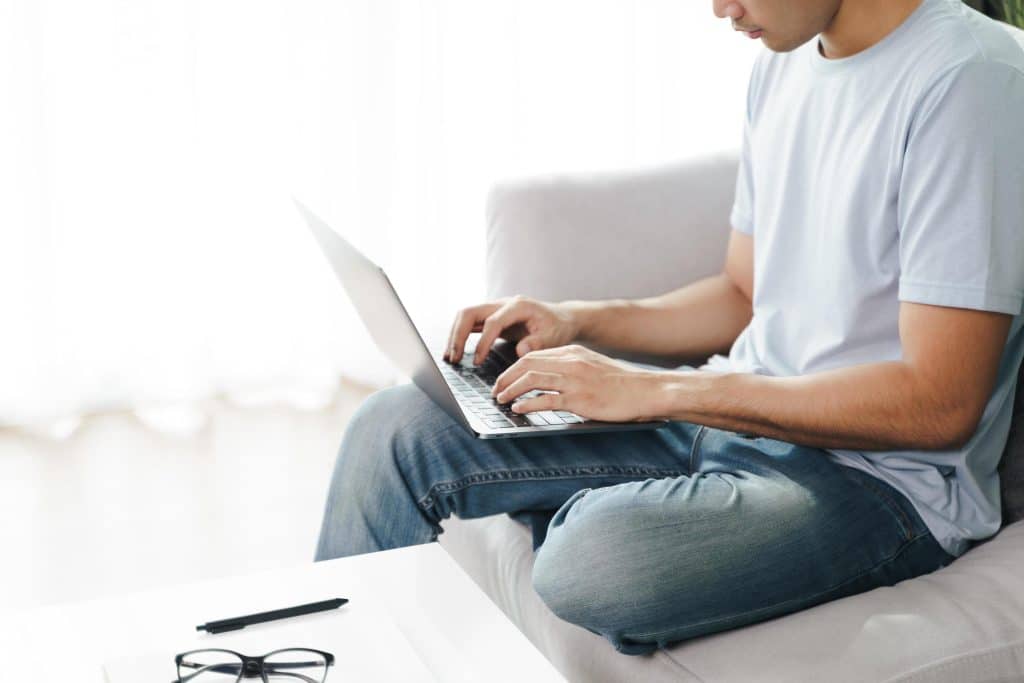 "Homem trabalhando remotamente de casa, digitando em seu notebook enquanto está sentado no sofá.