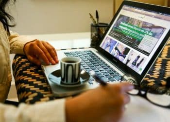 Mulher trabalhando remotamente em casa, digitando no laptop com xícara de café e caderno para ilustrar o conceito de teletrabalho.