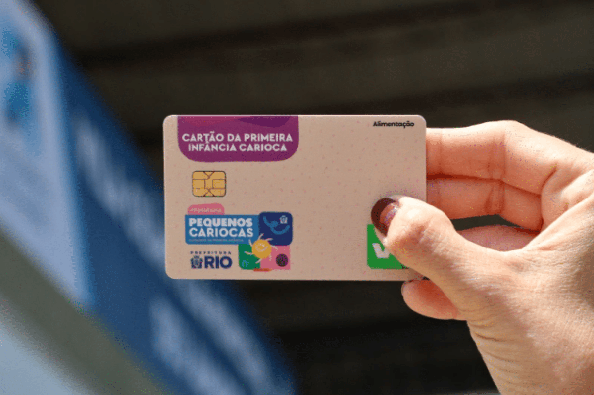 Mão segurando um cartão do programa Pequenos Cariocas, da Prefeitura do Rio, usado para benefícios de alimentação.