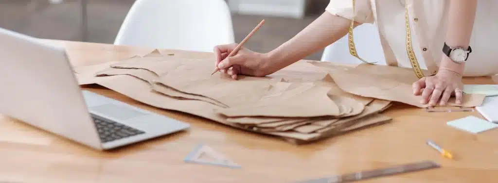 Detalhe de uma profissional de moda criando moldes de papel com lápis e régua, ao lado de um notebook.