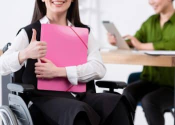 Mulher em cadeira de rodas segurando pasta rosa e fazendo sinal de positivo com a mão.