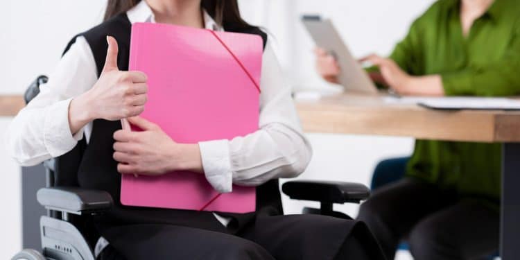 Mulher em cadeira de rodas segurando pasta rosa e fazendo sinal de positivo com a mão.