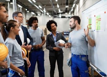 Equipe diversificada de técnicos, gestores e operários em reunião industrial com gráficos de desempenho ao fundo