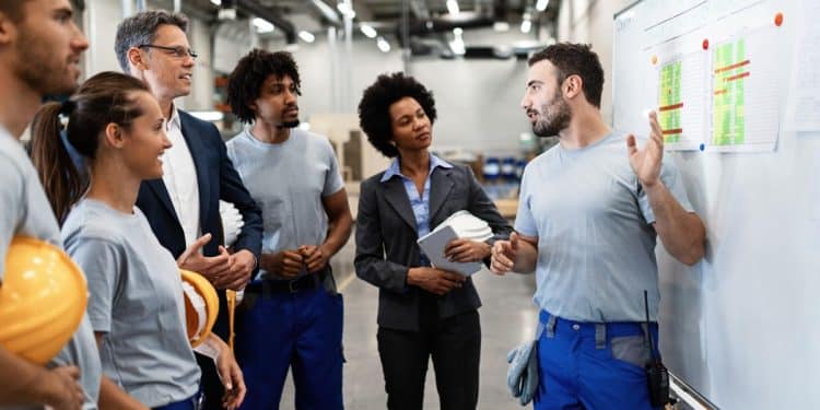Equipe diversificada de técnicos, gestores e operários em reunião industrial com gráficos de desempenho ao fundo