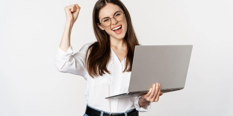 Mulher jovem e sorridente com óculos segurando um laptop cinza, fazendo gesto de comemoração com o punho.