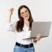 Mulher jovem e sorridente com óculos segurando um laptop cinza, fazendo gesto de comemoração com o punho.
