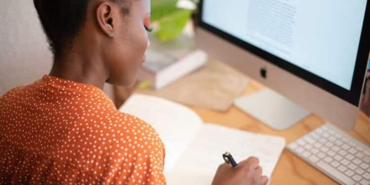 Jovem profissional independente trabalhando em home office, anotando ideias em caderno enquanto visualiza texto em monitor.