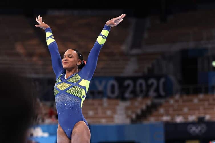Ginasta brasileira vestindo uniforme azul e amarelo com braços erguidos durante competição nas Olimpíadas Tóquio 2020