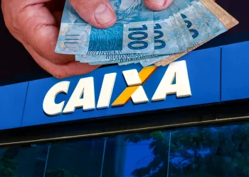 Mão segurando cédulas de dinheiro reais em frente a agência da Caixa Econômica Federal