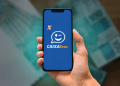 Mão segurando um smartphone com o aplicativo Caixa Tem aberto, mostrando o logotipo e um ícone de "piscadinha" sobre um fundo de notas de R$ 100 desfocadas, simbolizando o pagamento e a facilidade de acesso.