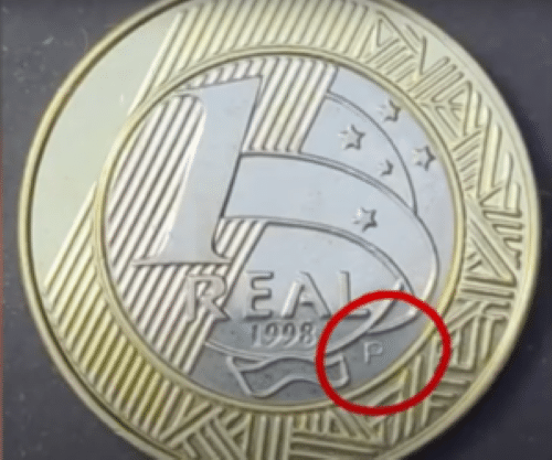 Close-up da moeda de 1 Real de 1998, com a letra "P" de "Prova" circulada em vermelho. Esta marca é o segredo para identificar a variante rara que pode valer até R$ 30 mil.