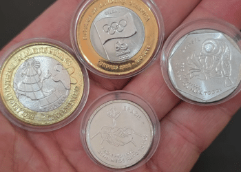 Mão segurando um conjunto de quatro moedas raras comemorativas do Real em cápsulas protetoras, incluindo modelos das séries FAO (mãos e lavrador) e Bandeira Olímpica. A imagem ilustra o potencial de valorização de coleções, que pode alcançar R$ 890 com a peça mais rara.