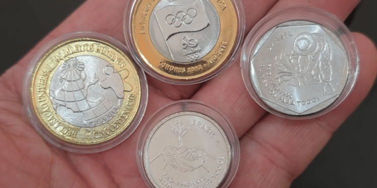 Mão segurando um conjunto de quatro moedas raras comemorativas do Real em cápsulas protetoras, incluindo modelos das séries FAO (mãos e lavrador) e Bandeira Olímpica. A imagem ilustra o potencial de valorização de coleções, que pode alcançar R$ 890 com a peça mais rara.