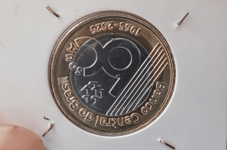 Moeda de 1 Real comemorativa dos 60 anos do Banco Central do Brasil (1965-2025) fixada em um holder, ideal para verificação de erros como o reverso invertido.